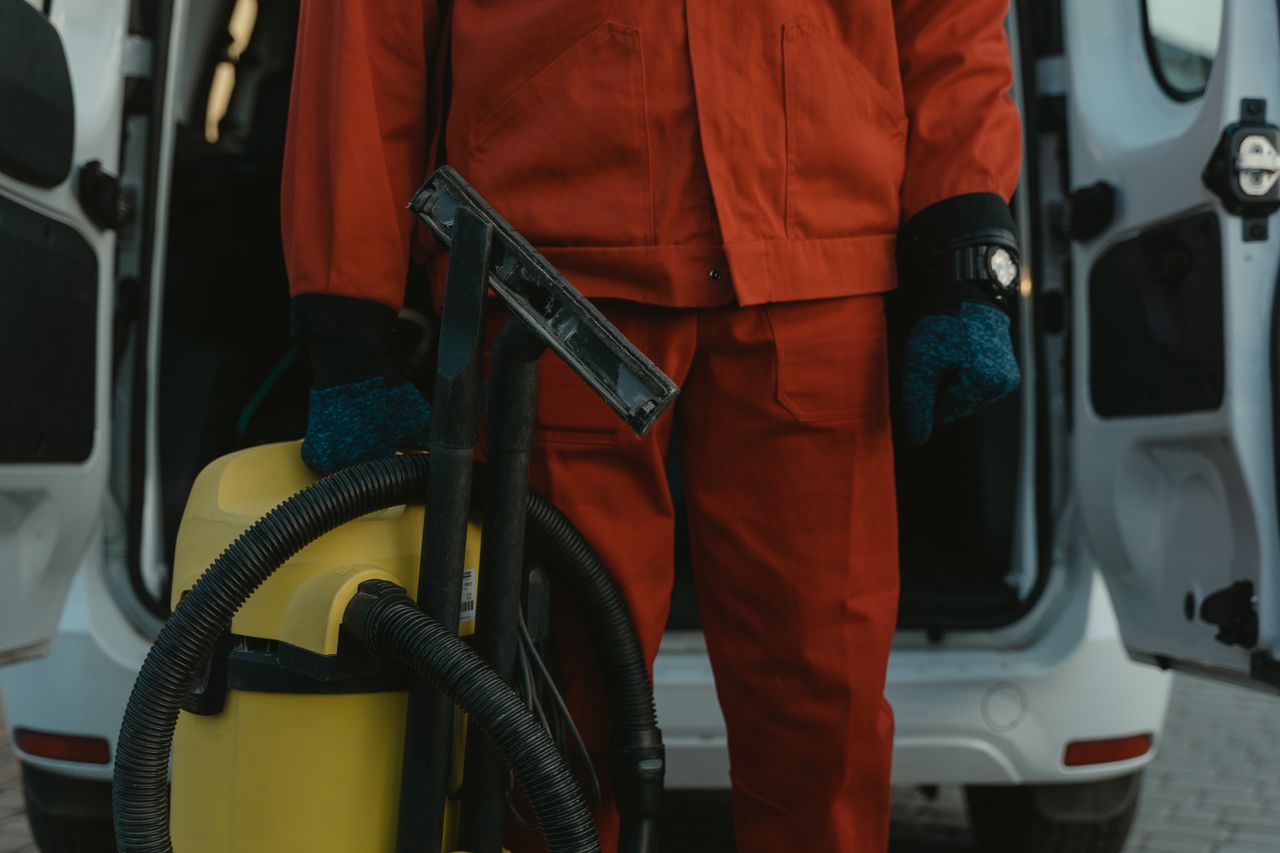Un professionnel du nettoyage en uniforme rouge tient un aspirateur près d’un van, prêt à intervenir.